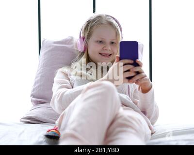 modern life of generation Z. teenage girl in pajamas and headphones in the room on the bed listens to music from a smartphone. Stock Photo