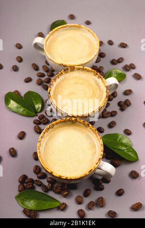 Three cups of coffee with foam among coffee beans and leaves on a gray table. Stock Photo