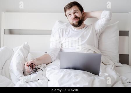 Blogger distracted from working with a laptop, took off glasses and looks up thoughtfully, sitting in bed. Creative process. Stock Photo