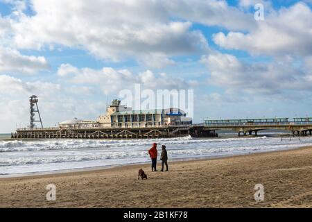 Couple walking along seashore with their dog at Bournemouth beach by Bournemouth pier at Bournemouth, Dorset UK on a sunny cold day in February Stock Photo