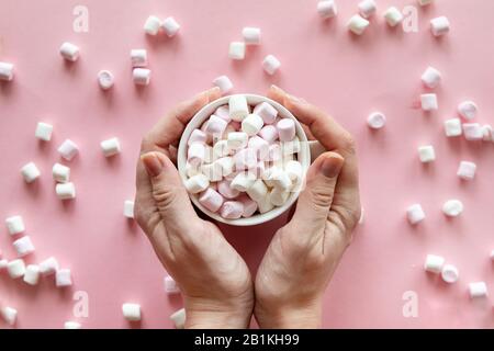Woman hand holding a cup with pink and white mini marshmallows on pink background Stock Photo