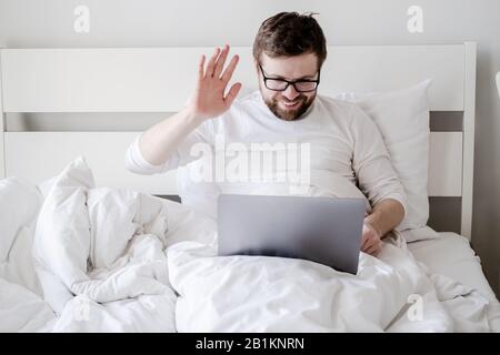 Man communicates remotely through a video call from a laptop, he looks at the screen, smiles and waves hand while sitting in bed. Stock Photo