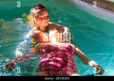 Beautiful senior woman receiving water massage in swimming pool from female therapist with sun rays falling on face in spa centre