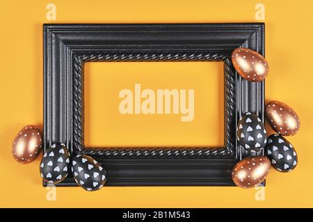 Easter design with blank black elegant picture frame surrounded by modern golden, black and white easter eggs on yellow background Stock Photo
