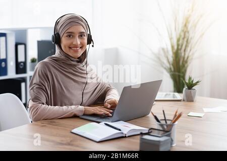 Arab female call center operator in hijab and headset at workplace Stock Photo