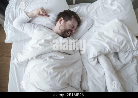 Man in white pajamas has a terrible nightmare, he grinds teeth and prepared a fist for protection, lying in his bed. Stock Photo