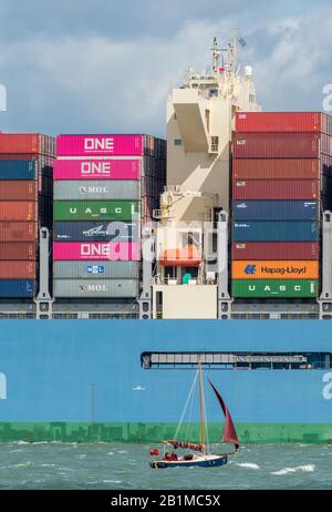 a large container carrying ship in the solent approaching the port of southampton docks with a small sailing yacht or dinghy next to it difference. Stock Photo