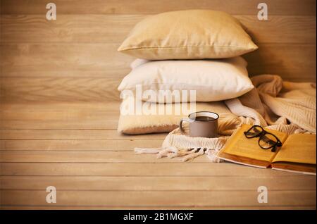 Grey mug of hot drink and eyeglasses on old opened textbook beside warm blanket and soft cushions on wooden bench at home Stock Photo