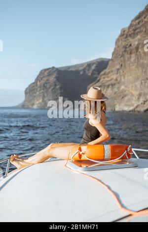 Woman enjoying ocean voyage sitting with lifebuoy on the yacht nose while sailing near the breathtaking rocky coast on a sunset. Concept of a luxury summer recreational pursuit and travel Stock Photo
