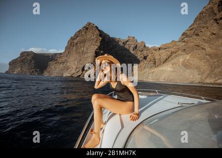 Woman in swimsuit and sun hat enjoying ocean voyage, sailing on a yacht near the breathtaking rocky coast on a sunset. Concept of a luxury summer recreational pursuit and travel Stock Photo