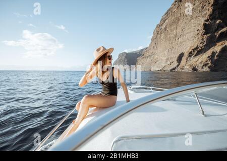 Woman in swimsuit and sun hat enjoying ocean voyage, sailing on a yacht near the breathtaking rocky coast on a sunset. Concept of a luxury summer recreational pursuit and travel Stock Photo