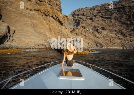 Woman in swimsuit and sun hat enjoying ocean voyage sitting on the yacht nose while sailing near the breathtaking rocky coast on a sunset. Wide seascape view Stock Photo