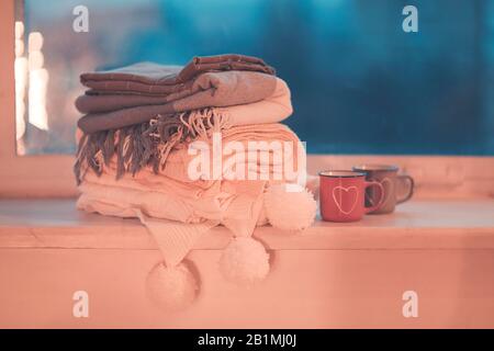 Stack of knitted plaids with gray and pink mugs on cozy window Stock Photo