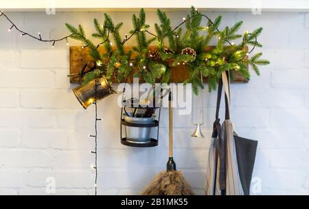 Old fashioned home utensils and apron hanging on wooden board decorated with spruce branches and light garland on white brick wall Stock Photo