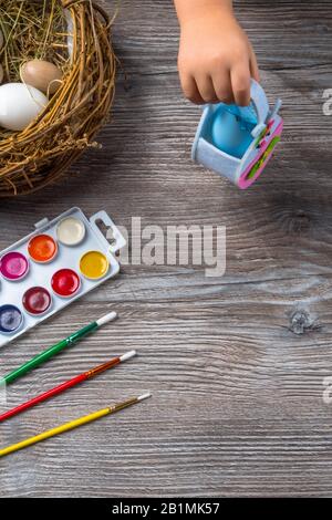 easter eggs in nest and colorful paints and brushes on wooden background. Baby beginning to painting. Close up Stock Photo