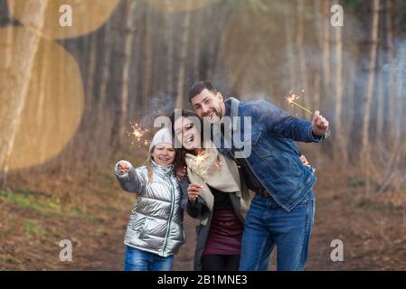 Cheerful young family with daughter dressed in warm clothes with sparklers in hands embracing and looking at camera while standing against forest back Stock Photo