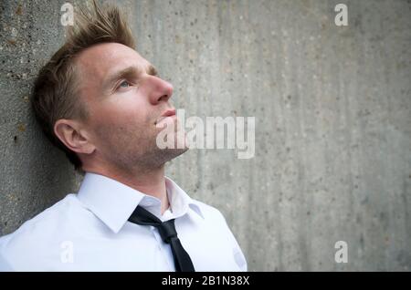 Serious man in white shirt and skinny black tie leaning against a textured concrete wall looking up Stock Photo