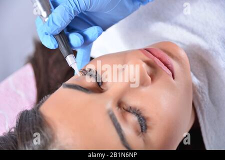 Cosmetologist applying permanent makeup on eyes on the beautiful girl in beauty studio. Eyeliner tattoo with a professional tool. Close up Stock Photo