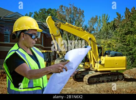 Female, Latino, black, Native American reading plans on construction site in front of site and bulldozers Stock Photo