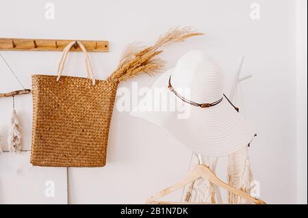 A white straw women's hat with wooden beads on a white hanger against a white wall and a straw brown bag with a bouquet of wheat and spikelets. Bohemi Stock Photo