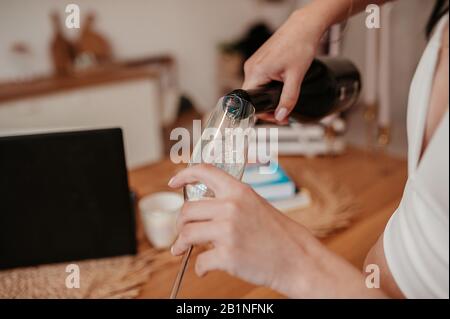 a woman's light-skinned hand holds a green glass bottle and pours a drink from it into a beautiful glass champagne glass in a white modern interior, w Stock Photo