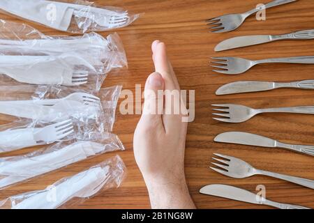 Plastic versus reusable cutlery concept: Man hand between disposable plastic sets and a stainless steel fork / knife set, on a walnut table top. Stock Photo