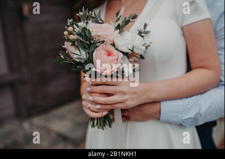 man hard hugs a woman in a white dress from behind and together they hold a chic elegant bouquet of brides from pink peony and white roses Stock Photo