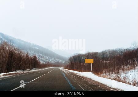 empty asphalt road with markings and empty yellow rectangular road sign on a background of foggy snow hills and mountains Stock Photo