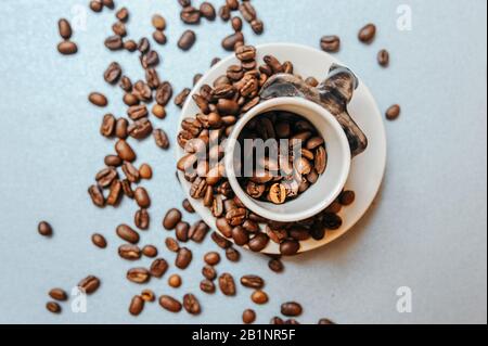 white mug full of coffee beans is standing on a white plate and grains are scattered around Stock Photo