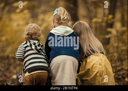 three friends of children, two girls and one little boy, are very stylishly dressed and stand with their backs in the forest, the children keep a secr Stock Photo