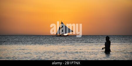 (Selective focus) Stunning view of a boat sailing during a beautiful sunset in the background and the silhouette of a blurred person in the foreground. Stock Photo