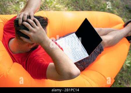 Worried shoked young man holding head like big problem and looking on laptop in outdoor. Stock Photo
