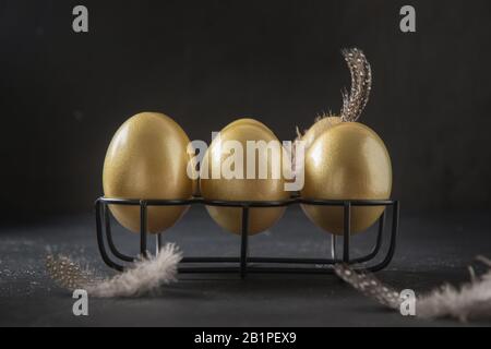 Chicken golden eggs in stand decorated feathers on black background. Easter dark concept. Close up. Stock Photo