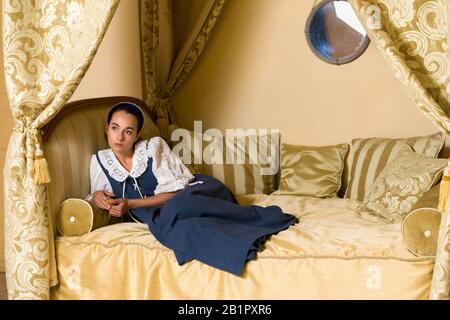 Dutch Master style portrait of a beautiful young woman in authentic renaissance costume lying on a luxury antique canopy bed in a golden bedroom of a Stock Photo