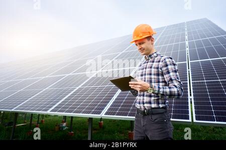 Worker in a helmet carries out work in a tablet on the background of a large station of solar panels. Photovoltaic power supply systems. Green energy concept design. Stock Photo