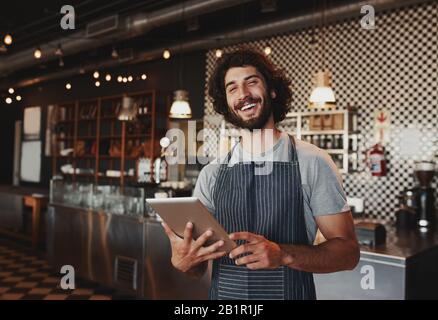 Handsome young caucasian coffee shop owner wearing apron laughing using digital tablet Stock Photo