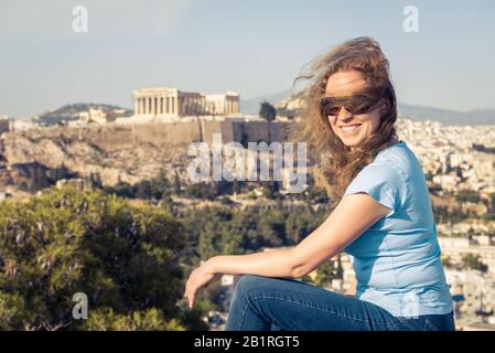 Young pretty woman sits at hilltop in Athens, Greece. Beautiful adult girl tourist smiles overlooking Athens city center. Attractive happy person agai Stock Photo