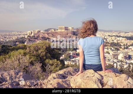 Young woman looks at cityscape of Athens, Greece. Adult girl tourist relaxes on hill top overlooking Acropolis of Athens in summer. Scenic view of Ath Stock Photo