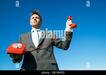 Serious businessman holding up the receiver of an old-fashioned red rotary dial telephone outdoors in sunny blue sky Stock Photo