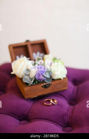 Two golden wedding rings on violet pillow near box with roses.