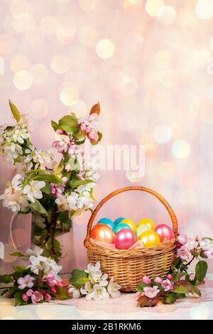 Easter festive composition of basket of painted Easter eggs, Apple tree twigs in glass decanter and on table with backdrop with hexagonal bokeh. Easte Stock Photo