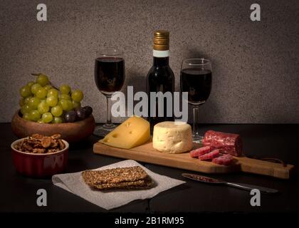Brie and swiss cheese on black stone cheese board, accompanied by fruit, nuts, crackers and two glasses of red wine, against light color background Stock Photo