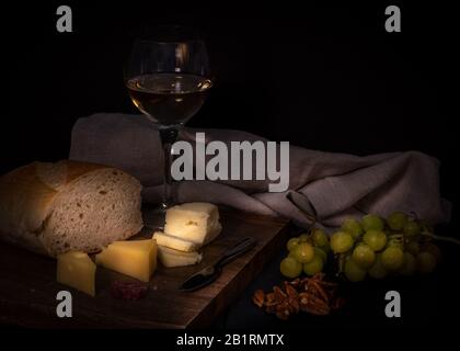 Brie and swiss cheese onwooden cheese board, accompanied by fruit, nuts, crackers and one glass of white wine, against black color background Stock Photo