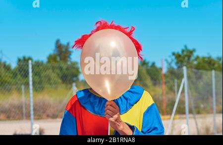 closeup of a scary clown wearing a colorful yellow, red and blue costume outdoors, holding a balloon in front of his face Stock Photo