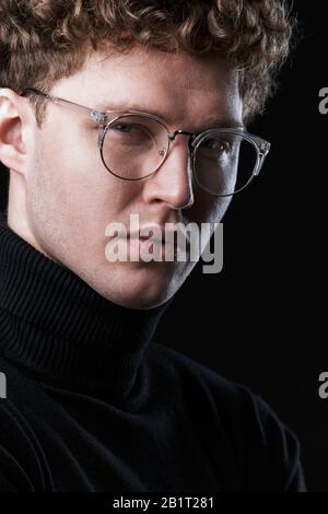Close up of a handsome confident young curly haired businessman wearing turtleneck standing infront of isolated black background, posing in glasses Stock Photo