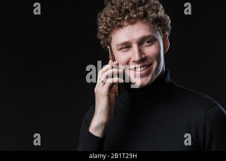 Portrait of a successful cheerful young attractive curly haired businessman standing infront of black background, talking on mobile phone Stock Photo