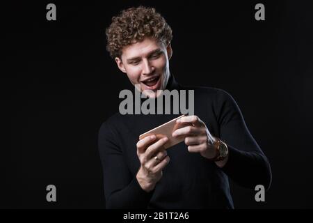 Portrait of a successful cheerful young attractive curly haired businessman standing infront of black background, playing on mobile phone Stock Photo