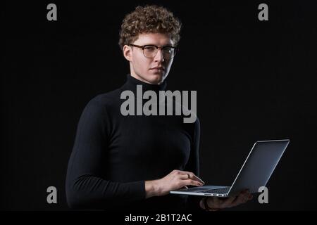 Handsome confident young curly haired businessman wearing turtleneck standing infront of isolated background, working on laptop computer Stock Photo