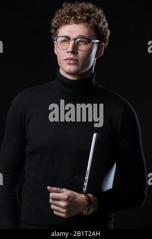 Handsome confident young curly haired businessman wearing turtleneck standing infront of isolated background, carrying laptop computer Stock Photo