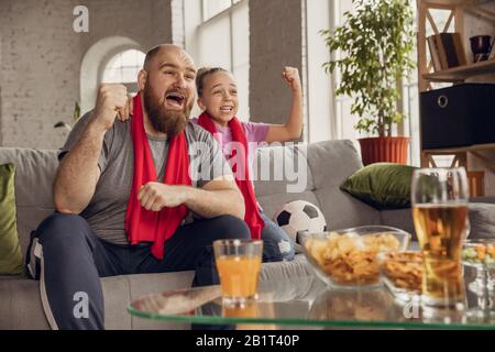 Excited, happy daughter and father watching football, soccer match on the couch at home. Fans emotional cheering for favourite national team. Having fun together, supporting. Sport, TV, championship. Stock Photo